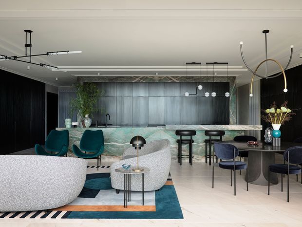 A refined open-plan living and entertaining area with a striking marble-look kitchen island and bar, dark ribbed wall panelling, sculptural pendant lighting, curved upholstered seating, and a dining table. The space combines deep teal, black, pale stone, and warm timber accents in a contemporary, luxe interior.