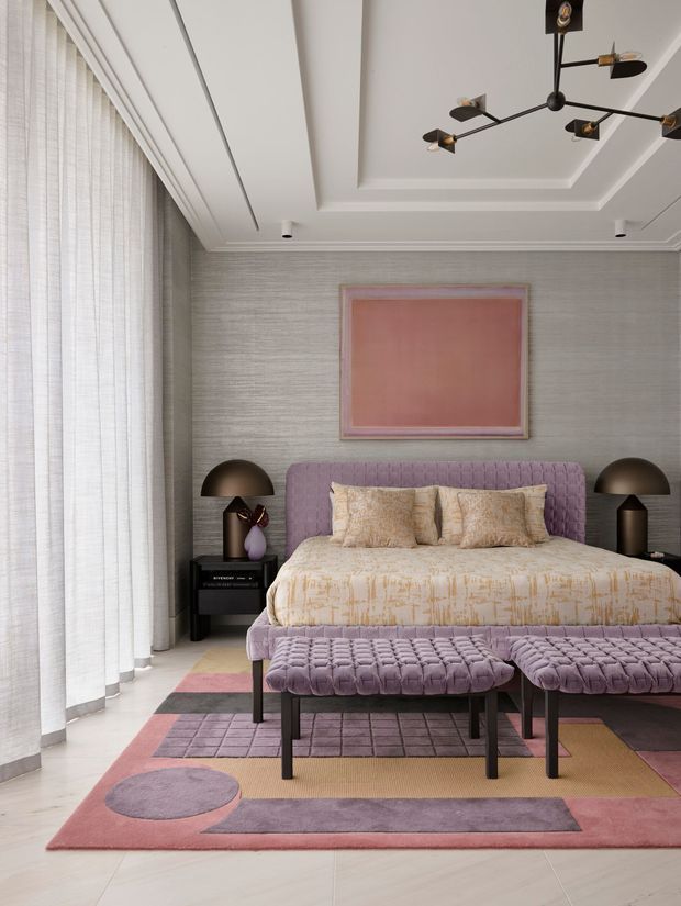 A refined bedroom with a large upholstered bed in muted lavender, matching tufted benches at the foot, and symmetrical dark bedside tables with dome lamps. Floor-to-ceiling sheer curtains line the left wall, while textured grey wall covering, layered tray ceiling detailing, and a contemporary sculptural chandelier add architectural interest. A large pink artwork and a geometric pink, lavender, and taupe rug create a soft, coordinated palette.
