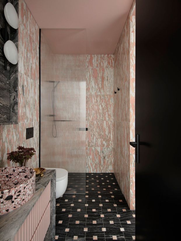 A compact, highly designed bathroom viewed from the doorway. The room features pink-and-white veined wall tiles, a dark textured tiled floor with small pale square inlays, a terrazzo-style pink vanity basin and benchtop, a white toilet, a black-framed fluted glass shower screen, dark marble-look wall surfaces, and black tapware, lighting, switches, and door hardware. The palette is bold pink, black, white, and grey with a luxurious, dramatic feel.