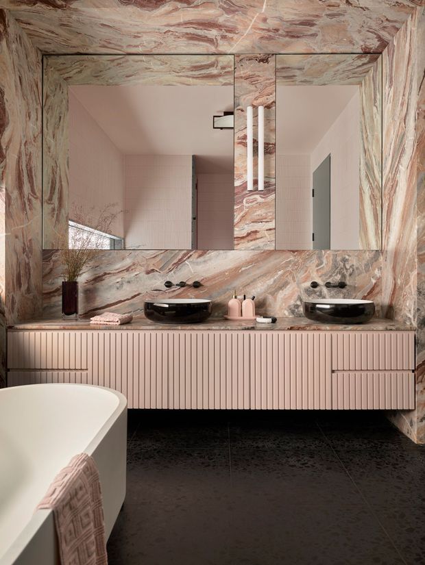 A luxurious contemporary bathroom featuring a double vanity with black vessel basins, ribbed blush-pink cabinetry, and a large frameless mirror. Dramatic pink and cream veined stone clads the walls, ceiling, vanity benchtop, and mirror surround. A freestanding white bathtub with a textured pink towel occupies the foreground, while pale pink wall tiles and dark floor tiles complete the refined, tonal interior.
