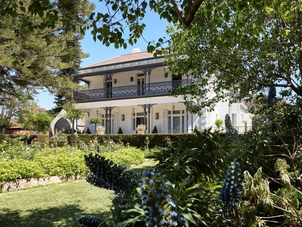 A grand two-storey heritage-style residence viewed through a mature landscaped garden. The light-coloured rendered facade features a red pitched roof, full-width upper-level verandah with ornate dark metal balustrades, decorative columns and brackets, tall multi-pane windows, and formal garden planting in the foreground.
