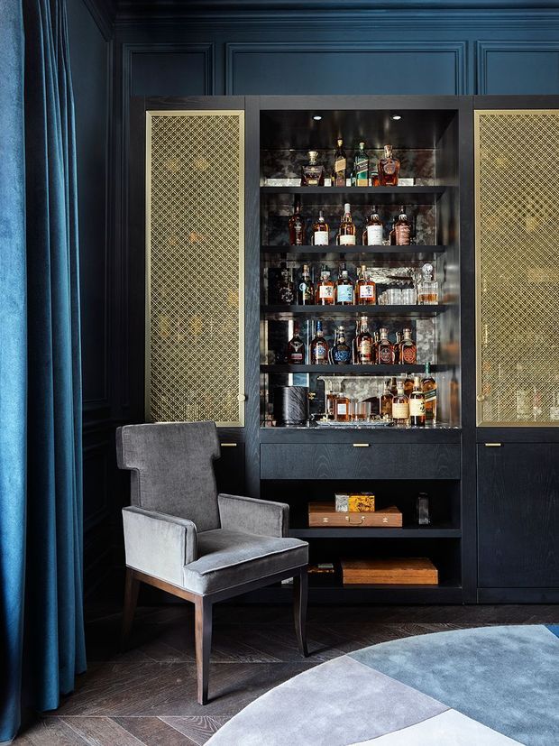 A moody, richly detailed home bar with dark blue-black paneled walls and built-in cabinetry. The central open shelving displays a collection of liquor bottles and glassware, flanked by doors with gold-patterned mesh or lattice inserts. A grey upholstered armchair sits beside the joinery, with a blue curtain at left and a partially visible geometric area rug over dark timber flooring.