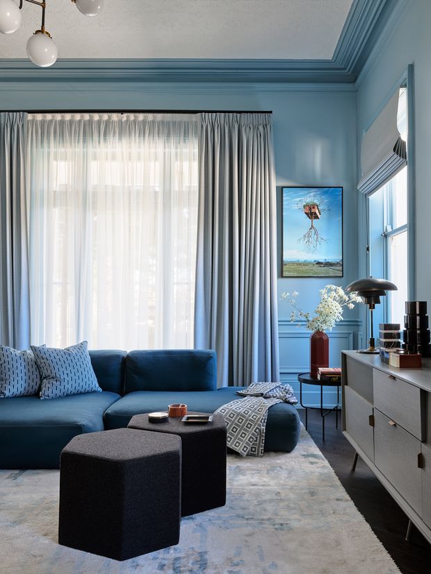 A refined living room with a deep blue sectional sofa, patterned cushions, dark ottomans, a pale area rug, and a streamlined mid-century-style sideboard. The room features blue painted walls with substantial decorative cornices and wall panelling, tall layered curtains, a textured decorative ceiling, framed artwork, and sculptural pendant and table lighting. The overall scheme is cool, elegant, and cohesive, with strong blue tones and restrained geometric detailing.