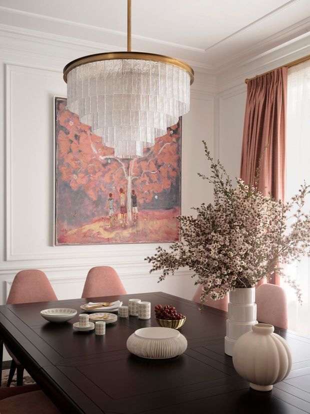 Elegant, traditionally detailed dining room with a dark rectangular dining table, blush upholstered dining chairs, white decorative wall panelling, a large abstract floral painting, and a dramatic tiered glass pendant light with a brass-toned rim. Soft pink drapery and abundant floral branches create a refined, warm, decorative setting. Tableware and sculptural white ceramic vessels are arranged as styling accents.