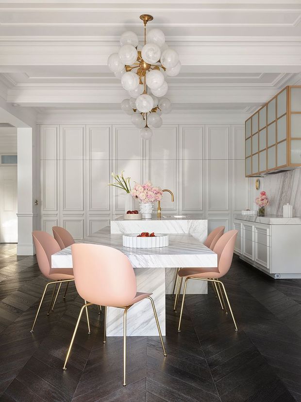 A refined, bright dining area within an open-plan kitchen, featuring a marble dining table with a sculptural pedestal base, blush-pink upholstered dining chairs with gold-toned legs, white paneled walls and cabinetry, a marble island and benchtops, and a striking multi-globe brass pendant light. Dark herringbone timber flooring creates contrast, while a brass-framed glazed cabinet and floral arrangements add decorative detail.