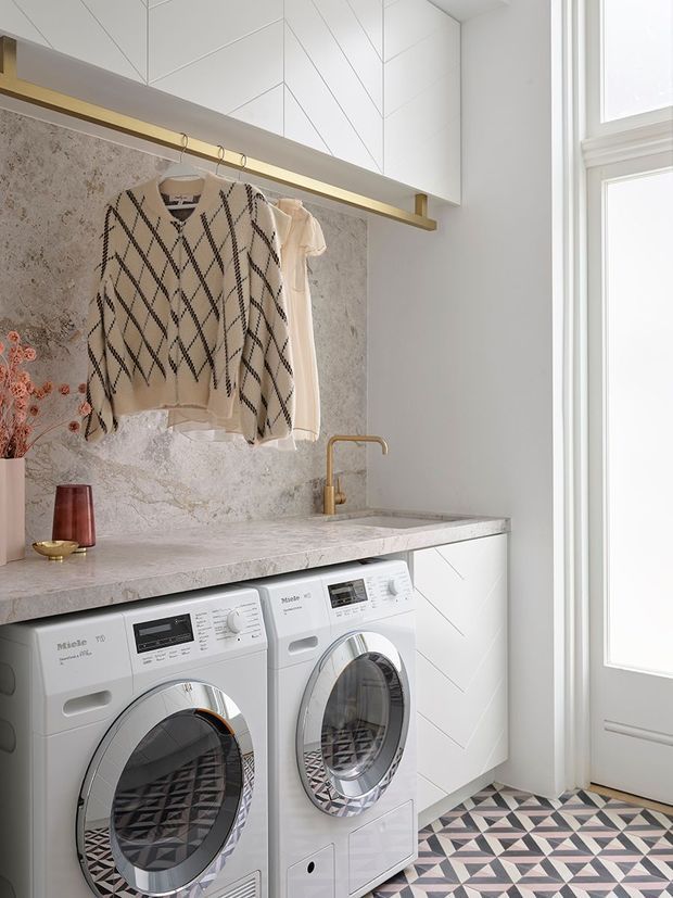 A bright, refined laundry room with white cabinetry, integrated washing machine and dryer, a pale stone countertop and backsplash, and a brass-toned tap. A hanging rail displays clothing beneath geometric white cabinetry, while patterned geometric floor tiles add contrast. Tall glazed doors or windows bring in abundant natural light.