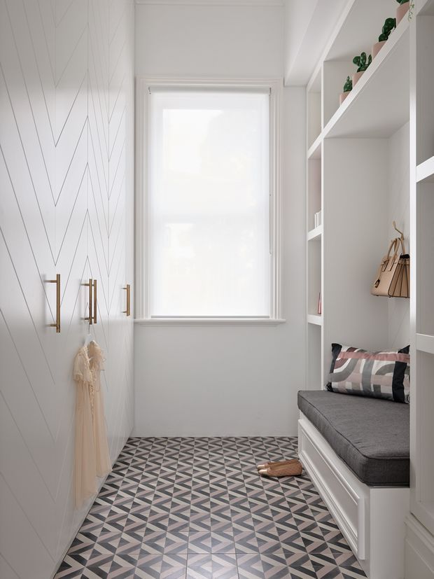 A narrow, bright walk-in wardrobe or dressing room with custom white built-in shelving and a window seat. The left wall features geometric line-patterned panelling with brass hooks and hanging garments, while open shelving on the right displays small plants and accessories above a grey upholstered bench. The floor is covered in a bold geometric patterned tile in grey, black, white, and muted pink tones. The space has a refined, contemporary built-in design with abundant natural light.