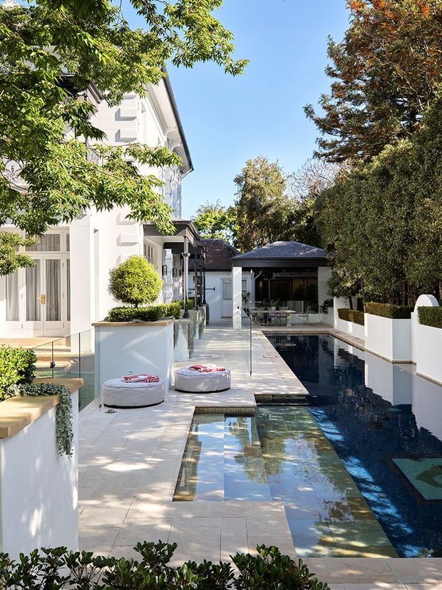 A bright, landscaped courtyard centered on a long narrow swimming pool beside a white multi-storey residence. Pale stone paving, white rendered planter walls, clipped hedges, mature trees, glass pool fencing, and a covered poolside pavilion create a refined indoor-outdoor setting. Two round cushioned outdoor loungers sit along the pool deck.