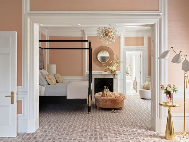 A refined bedroom viewed through a wide decorative opening, featuring a black four-poster bed, pale bedding, blush-pink upholstered walls, elaborate white wall mouldings and trim, a patterned carpet, and a classic fireplace with mirror above. A tufted round ottoman, sculptural pendant light, floor lamp, and small floral arrangement add elegant decorative detail. An adjoining bright sitting or dressing area is visible beyond the bedroom.