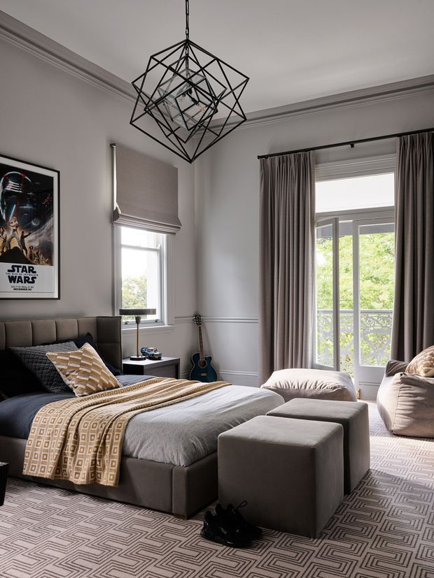 A refined bedroom with a grey upholstered bed, layered neutral bedding, geometric patterned rug, matching ottomans, and a large lounge chair. Grey curtains frame tall glazed doors opening to a balcony, while a sculptural black geometric pendant light, framed Star Wars poster, guitar, and decorative cushions add personality. The room features traditional wall moulding and cornice details combined with contemporary furnishings.