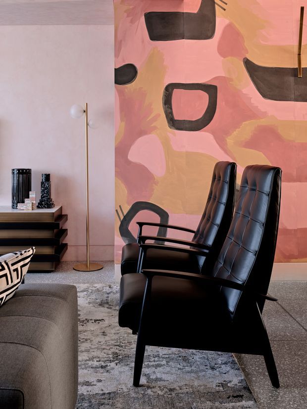 A stylish living room vignette featuring two black leather high-back lounge chairs in the foreground, a grey upholstered sofa edge, a patterned grey rug, and a dark timber media or storage console. A large abstract pink, coral, peach, and black artwork forms a dramatic feature wall, complemented by a slim brass floor lamp and sculptural black decor. The space has a refined mid-century-inspired aesthetic with strong graphic contrasts.