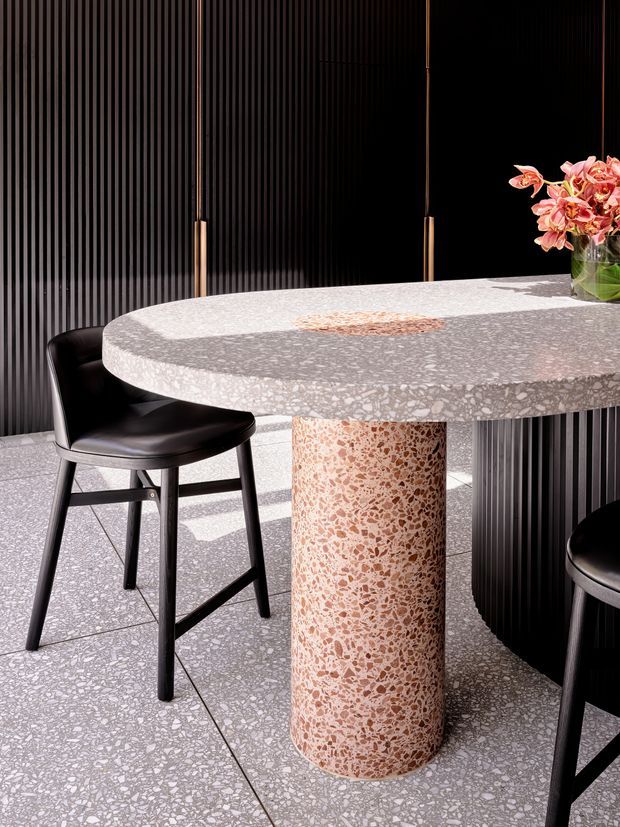 Close-up view of a refined dining area featuring a rounded terrazzo table with a thick top and cylindrical terrazzo pedestal base. The pale grey-white terrazzo has prominent warm peach and pink aggregate, coordinated with the terrazzo flooring. Black leather bar stools, a dark vertical fluted wall, slim brass-toned pendant lights, and a vase of pink flowers complete the composition. Strong natural light creates crisp shadows and a high-contrast, contemporary atmosphere.