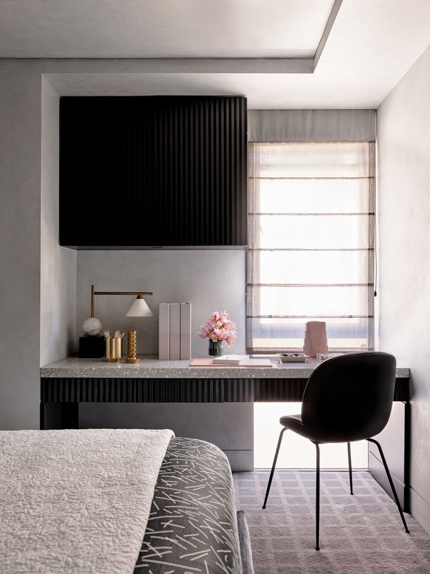 A refined bedroom with an integrated home office. A black fluted built-in wall unit and desk run beneath a large window with sheer Roman blinds. The room features soft grey plaster walls, a speckled stone-look desktop, black furniture and lighting, a patterned grey rug, and a bed in the foreground with textured white and grey bedding. The overall composition is calm, contemporary, and monochromatic with warm brass accents and soft natural light.