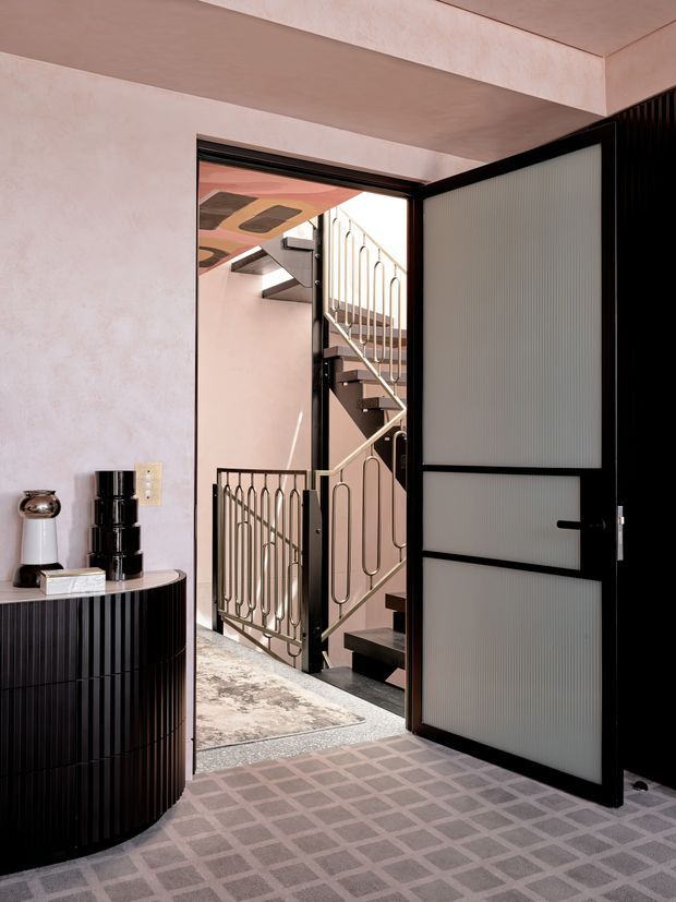 A refined interior entryway with a partially open black-framed door featuring translucent ribbed glass panels. The space has softly textured blush-pink plaster walls, a dark fluted curved console with decorative objects, and a patterned grey-and-pink carpet. Beyond the doorway is a stairwell with dark treads and distinctive sculptural metal balustrades, plus a patterned ceiling detail.