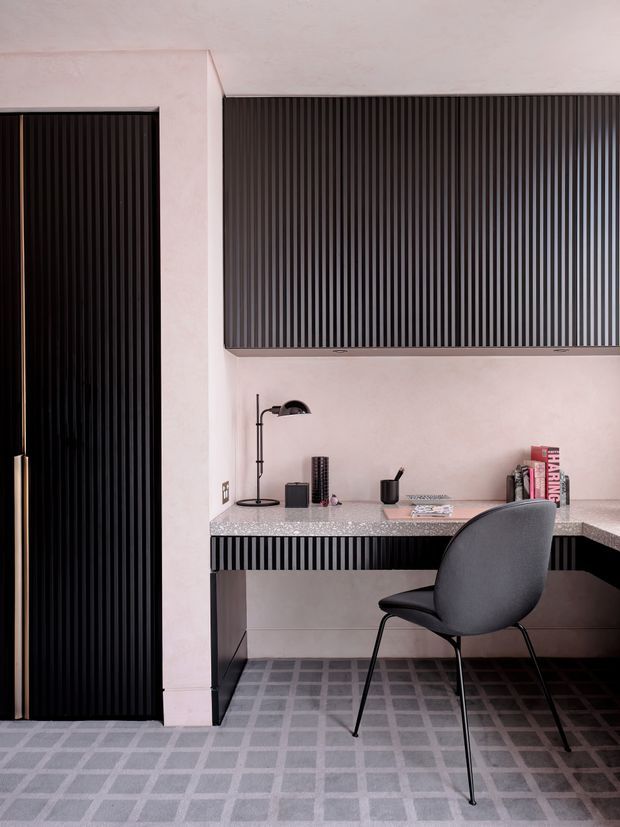 A compact, refined home office with a built-in desk and extensive black vertically ribbed cabinetry. The room features pale pink plaster walls, a terrazzo-style benchtop, dark cabinetry, a black desk lamp, and a grey upholstered chair. The floor is finished with a grey grid-patterned carpet or tile-like surface, creating a strong geometric, monochromatic look.