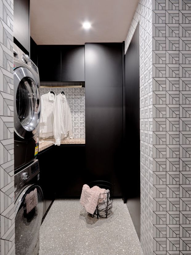 Compact contemporary laundry room with stacked front-loading washer and dryer on the left, tall matte-black cabinetry and storage along the back wall, and a hanging rail with white garments above a stone-look counter. The walls are clad in a bold geometric patterned tile, while the floor has a light grey terrazzo-like finish. A small wire laundry basket with folded pale pink textiles sits in the centre.