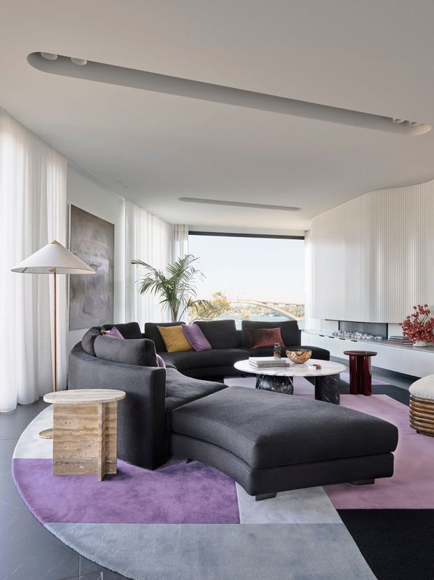 A bright, sophisticated contemporary living room with floor-to-ceiling sheer curtains, expansive glazing, a large curved dark sectional sofa and chaise, and a sculptural layered area rug in purple, grey, and black. A white marble coffee table, sculptural side tables, indoor palm, abstract artwork, and a fluted white built-in media wall complete the refined interior. Linear recessed ceiling lighting and a distant coastal-looking view are visible.