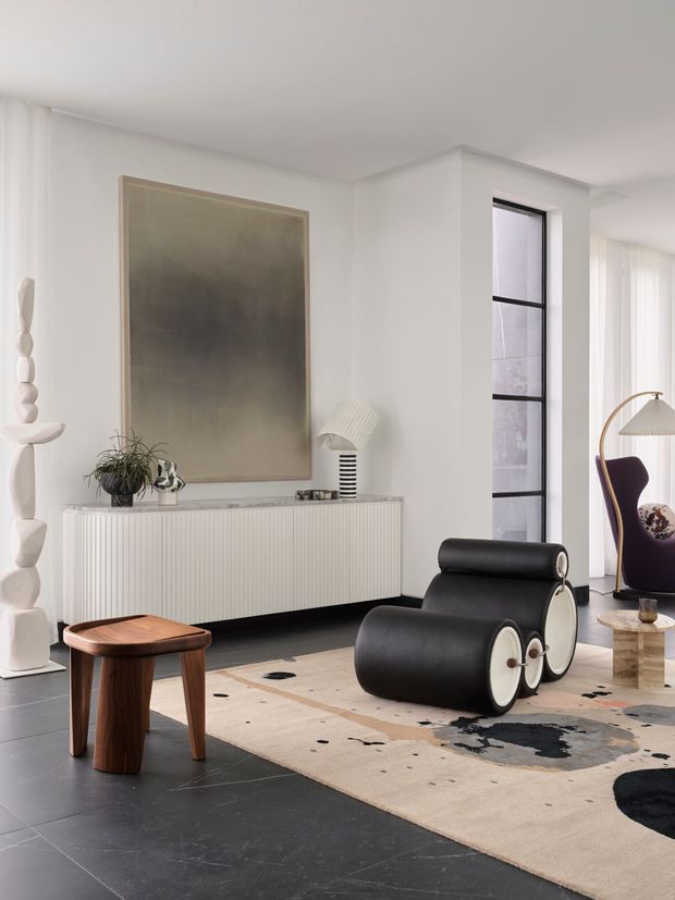 A refined contemporary living room with a predominantly white interior, black tiled flooring, and a large neutral area rug with abstract dark and terracotta accents. The room features a sculptural black leather lounge chair, a fluted white console with stone-look top, a large muted artwork, a small timber side table, and a glimpse of a curved seating area through the opening at right. Tall sheer curtains and slim black-framed windows provide soft natural light.