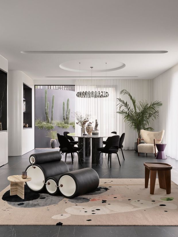 A refined open-plan dining and living space with a round black dining table and sculptural black chairs, set beside full-height sheer curtains and a large indoor palm. The room features dark stone-look flooring, a pale patterned rug, sculptural black-and-white lounge seating, a textured cream armchair, built-in display shelving, ribbed wall detailing, and contemporary decorative lighting. A courtyard-like planted view is visible through the glazing.