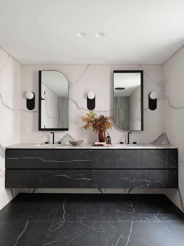 A refined contemporary bathroom featuring a double floating vanity with dark marble-look cabinetry and benchtop, large-format white marble-look wall surfaces, and matching dark veined floor tiles. Two black-framed mirrors, black wall sconces, and twin black tapware create a high-contrast, luxurious scheme. A small vase and basin accessories add warmth to the otherwise monochromatic space.