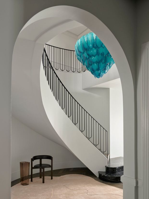 A refined, sculptural stairway viewed through a large white arched opening. The curved staircase has a black metal balustrade with looping vertical details, white fluted wall panelling, dark stone stair treads, and a striking turquoise sculptural pendant light suspended in the stair void. A small dark timber chair and side table sit on a pale rounded rug in the foreground.