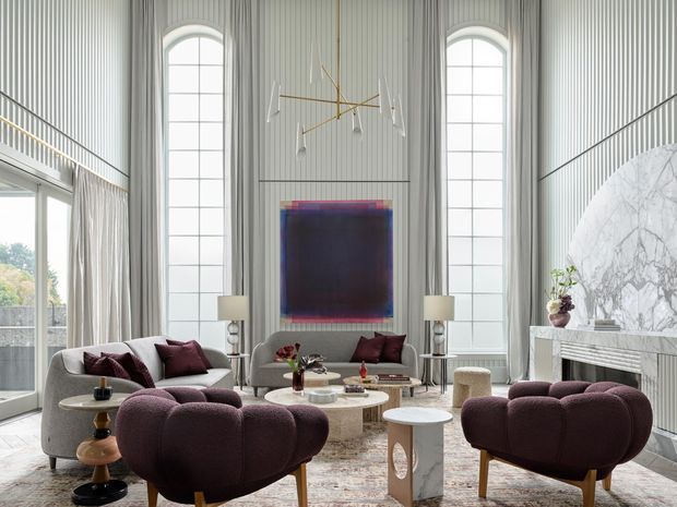 A sophisticated double-height living room with tall arched windows, floor-to-ceiling fluted wall panelling, a sculptural marble fireplace, and a layered seating arrangement in muted burgundy, grey, and cream. A large abstract artwork, contemporary chandelier, patterned rug, and sculptural side tables complete the refined interior.