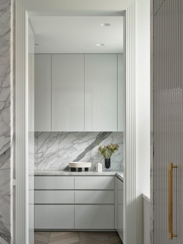 A refined, minimalist kitchen or kitchenette viewed through a fluted white architectural opening. The space features handleless pale cabinetry, a dramatic veined marble splashback and benchtop, integrated recessed ceiling lights, and dark herringbone timber flooring. A small counter stool, vase, and floral arrangement add restrained styling. A fluted-glass or ribbed door with a brass handle is visible at right.