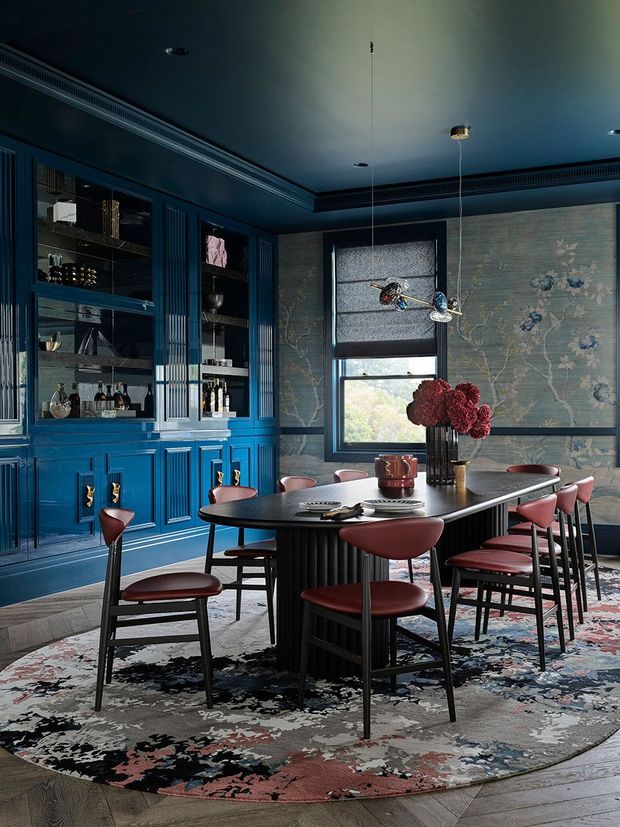 A richly styled dining room with a long dark dining table, sculptural black fluted base, and burgundy upholstered chairs. Deep blue built-in cabinetry with glass-front display storage lines the left wall, while botanical-patterned wallpaper, dark blue trim, a large patterned rug, and contemporary pendant lighting create a dramatic, refined atmosphere.