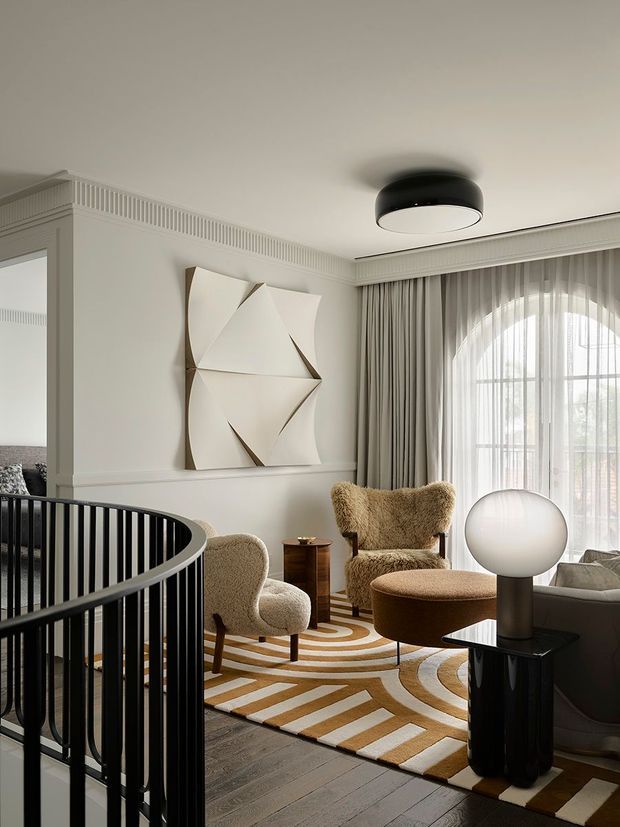 A refined sitting room with a curved black stair balustrade in the foreground, sculptural cream boucle lounge chairs, a rounded ottoman, a dark side table, and a large spherical lamp. The room features pale walls with decorative fluted cornice detailing, full-height drapery, an arched multi-pane window, geometric abstract wall art, dark timber flooring, and a bold cream, tan, and ochre geometric rug.