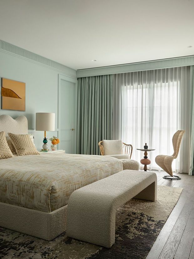 A refined bedroom with a large upholstered bed, textured neutral bedding, a curved upholstered bench, woven lounge chairs, and a small side table. Pale sage-green wall panelling and curtains frame full-height windows, while decorative ceiling moulding, layered sheers, and a muted patterned rug create a calm, elegant interior.