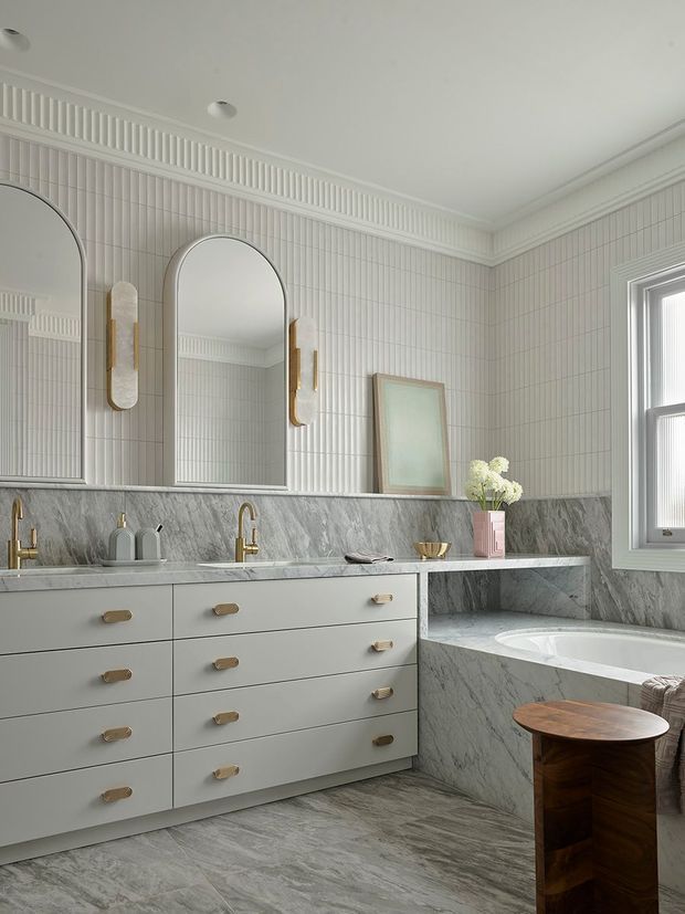 Elegant bathroom with a long double-sink vanity, pale grey cabinetry and brass hardware, marble-look wall and floor surfaces, a bathtub set into a marble-clad surround, and vertically grooved light wall tiles. Two arched mirrors with brass wall sconces sit above the vanity, while a large window provides natural light. A small dark timber stool adds contrast.