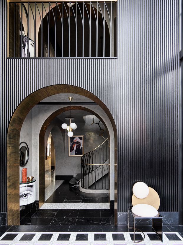 Dramatic interior view of a refined entry hall featuring tall ribbed dark wall panelling, layered rounded archways with dark metallic trim, black marble flooring with contrasting pale geometric inlays, a sculptural curved staircase with black balustrade, and statement pendant lighting. A sculptural chair and console are positioned at the right and left edges of the space. The composition is highly symmetrical and gallery-like with strong Art Deco-inspired detailing.
