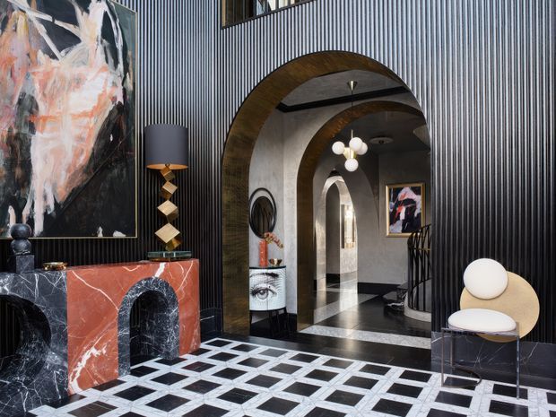 A dramatic, highly styled interior foyer or living space featuring black ribbed wall panelling, a large arched timber-trimmed opening, polished black-and-white geometric flooring, a veined red marble fireplace surround, sculptural lighting, contemporary artwork, and a sculptural lounge chair. The space has strong Art Deco influences with luxurious, high-contrast detailing and repeated arches leading into the hallway.