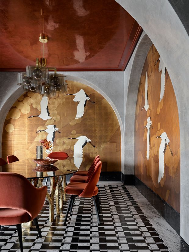 A dramatic, highly decorative dining room with a long glass dining table and sculptural red upholstered chairs. The room features arched plaster openings, a glossy copper-red ceiling, large gold-toned wall panels decorated with flying white cranes, a statement glass pendant chandelier, and a bold black-and-white checkerboard floor with marble-like veining. The overall aesthetic is richly layered, theatrical, and art-driven.
