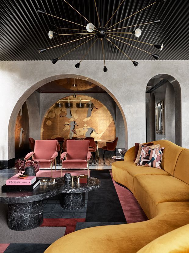 A richly layered living room with a mustard curved sectional sofa, pink velvet lounge chairs, and black marble coffee tables. Arched openings lead to a dining area with a dramatic metallic mural, while a sculptural multi-arm chandelier hangs from a dark ribbed ceiling. The space combines textured plaster walls, patterned flooring, saturated colours, and Art Deco-inspired detailing.