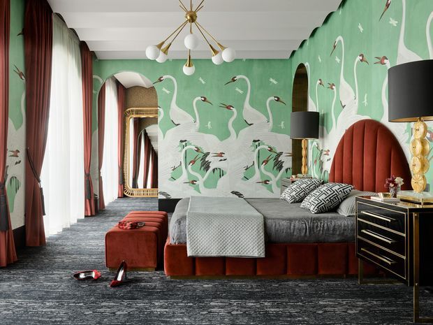 A richly decorated bedroom with a dramatic green flamingo-and-swan mural wallpaper, a large upholstered bed with a tall rust-red channel-tufted headboard, patterned cushions, velvet ottoman, dark textured carpet, floor-to-ceiling curtains, and ornate brass and black bedside furniture. A sculptural brass-and-white globe chandelier and mirrored arched detailing add a glamorous, eclectic character.