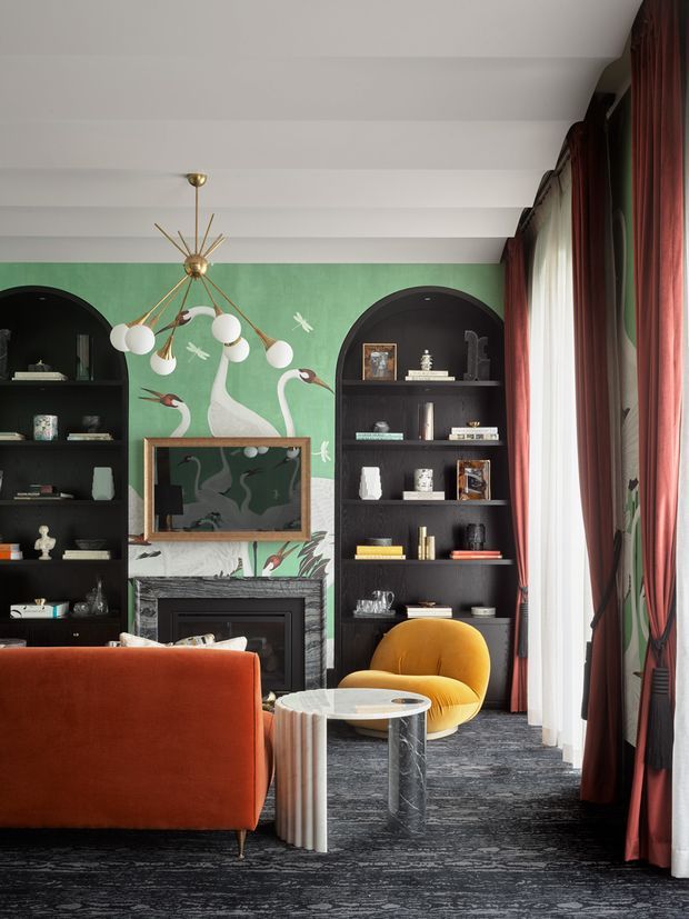 A richly styled living room with a dark patterned carpet, deep red-orange sofa, mustard-yellow lounge chair, and a small round marble coffee table. The room features a dramatic green botanical mural or wallpaper with painted birds, black arched built-in shelving flanking a dark fireplace, a framed artwork above the mantel, tall sheer and burgundy curtains, and a sculptural brass-and-glass chandelier. The overall look is eclectic, theatrical, and richly layered, with strong colour and pattern.