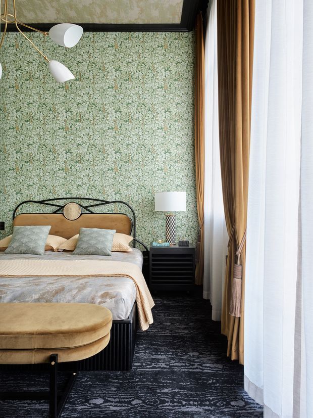 A richly layered bedroom featuring green botanical-patterned wallpaper, a black metal bed with a rounded upholstered headboard, patterned cushions, and a taupe upholstered bench at the foot. Dark patterned carpet, a black bedside cabinet, brass-toned curtains, sheer white drapes, and sculptural white pendant lighting create an elegant, vintage-inspired interior.