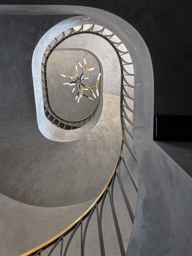 A dramatic upward view through a sculptural spiral staircase, showing curved grey plaster or concrete walls, dark metal balustrades with warm timber or brass-toned handrails, and a modern geometric pendant light suspended in the central void. The composition emphasises the looping stair geometry and vertical circulation space.