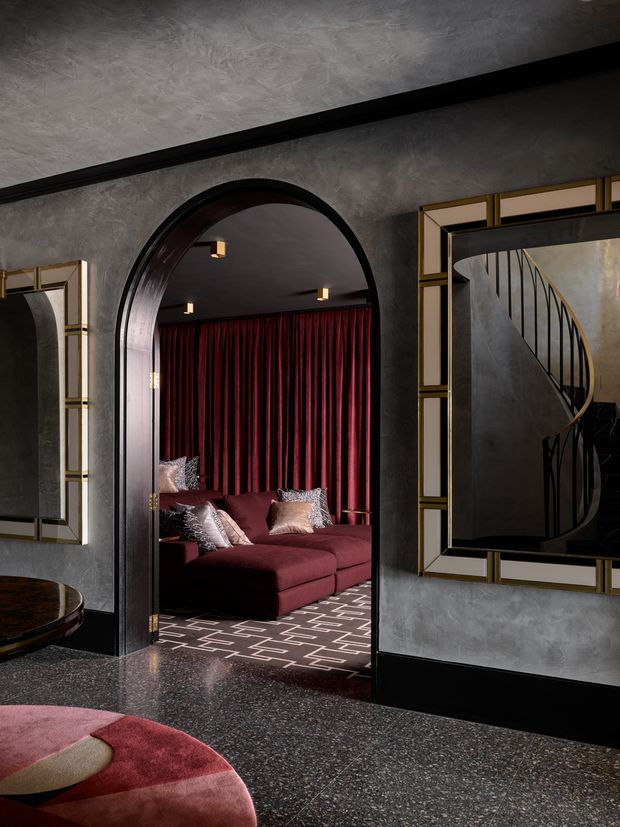A dark, luxurious interior hallway or sitting area with textured grey walls, black trim, and terrazzo-style flooring. A large arched doorway opens into a richly furnished lounge with a deep burgundy sectional sofa, matching floor-to-ceiling curtains, patterned rug, and decorative cushions. Geometric black-and-gold framed wall mirrors flank the doorway, with one reflecting a curved staircase. The overall design is dramatic and Art Deco-inspired, with rounded forms, metallic accents, and a moody jewel-toned palette.