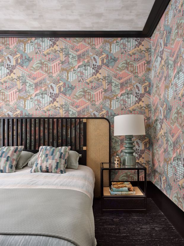 A richly patterned bedroom featuring full-height decorative wallpaper with pastel architectural and botanical motifs, a black vertical-slat and cane-panel headboard, layered patterned cushions, pale textured bedding, and a dark bedside table with a sculptural green lamp. Dark skirting, trim, and flooring create contrast, while the compact room has a polished, eclectic decorative character.