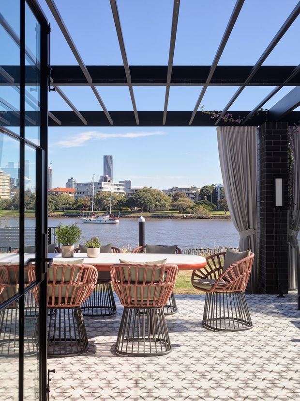 A bright waterfront outdoor dining terrace viewed from inside through large glazed doors. The terrace features a long dining table with woven coral-toned chairs, patterned pale tiled paving, a timber-and-metal pergola overhead, and a dark brick wall with curtains. Beyond the terrace is a river with a moored sailboat and an urban skyline.