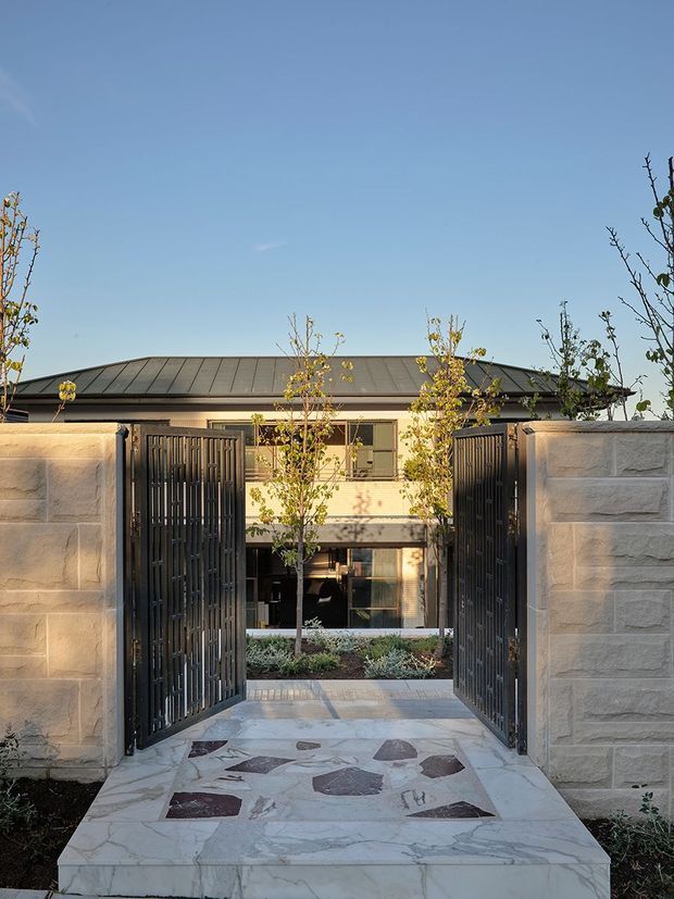 Front entry to a contemporary residence viewed through open black metal pedestrian gates. Pale stone masonry walls frame a patterned stone-paved path leading to a landscaped courtyard and a two-storey house with a low-pitched dark metal roof, rendered upper facade, glazing, and timber-toned detailing. Young trees and planting soften the structured entrance.