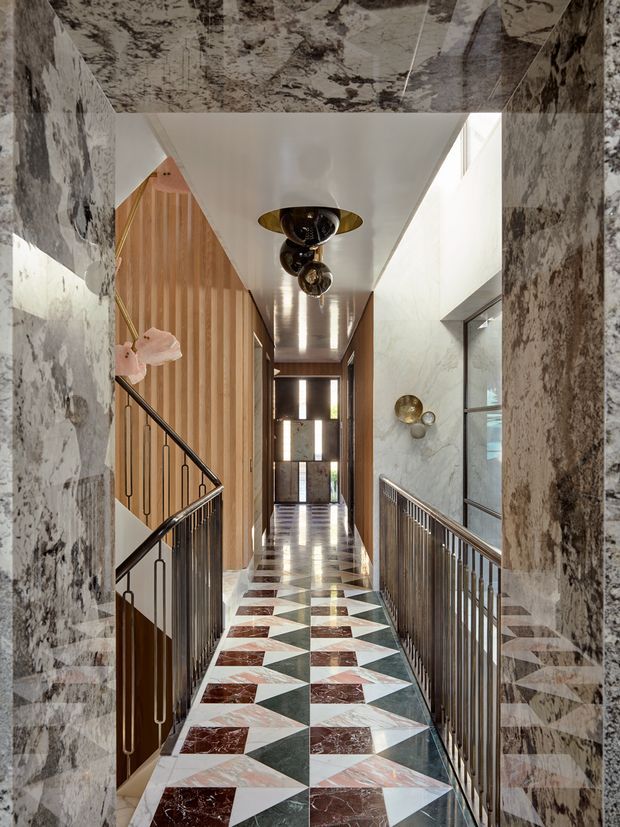 A dramatic, richly detailed interior hallway or landing viewed through a marble-clad opening. The corridor features a geometric patterned marble floor in white, green, and burgundy tones, matching veined marble wall and ceiling surfaces, warm vertical timber panelling, dark metal balustrades, sculptural brass-toned wall lights, and a decorative central pendant. Stair edges and railings indicate a split-level circulation space, with daylight entering from glazing at the far end and right side.