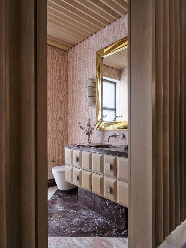 A compact, highly decorative bathroom viewed through a dark timber-framed opening. The room features a floating geometric timber vanity with sculptural drawer fronts, a dark burgundy-purple marble benchtop and floor, blush patterned wall covering, a brass-framed mirror, wall-mounted brass tapware, and a slatted timber ceiling. A toilet and window are visible in the background.