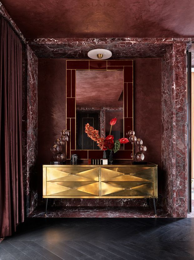 A dramatic, richly layered interior vignette featuring a sculptural gold geometric console with drawers, a large brass-trimmed rectangular mirror, and decorative lighting and floral arrangement. Deep burgundy plaster and veined marble frame the niche, with dark timber flooring and matching dark drapery creating a luxurious, moody Art Deco atmosphere.