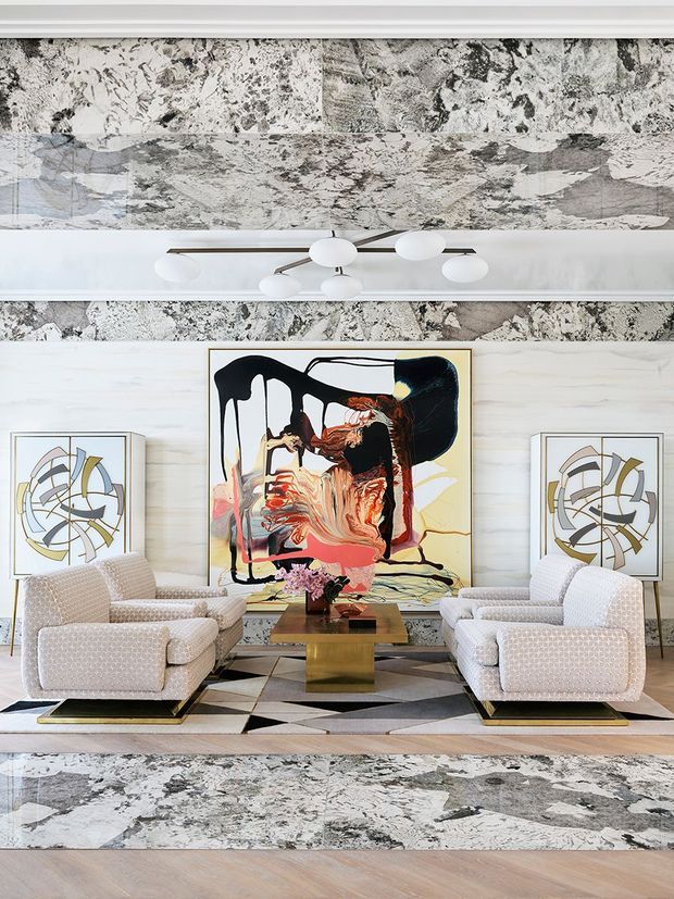 A refined living room with four upholstered armchairs arranged around a compact brass-toned square coffee table. The room features a dramatic abstract artwork as the focal point, flanked by two framed geometric artworks, with a sculptural multi-arm ceiling light. Large-format monochromatic marbled surfaces appear on the ceiling, upper wall bands and rug, contrasted with pale timber-toned wall panelling and flooring. The overall composition is symmetrical, polished and high-end.