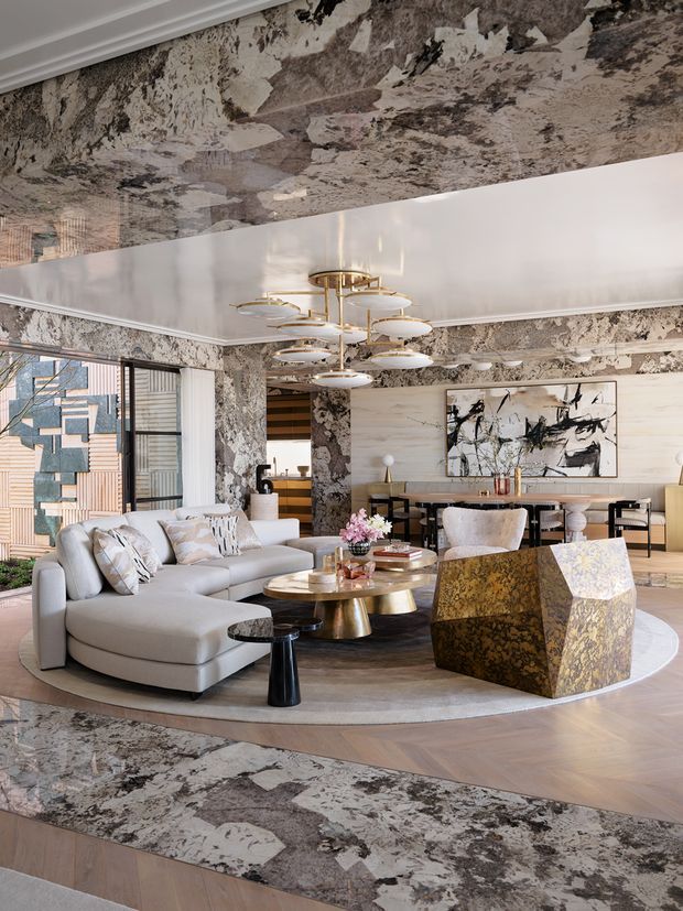 A sophisticated open-plan living and dining area featuring a large curved cream sectional, layered round coffee tables, sculptural metallic side tables, and a faceted gold-toned accent chair. Dramatic marble-patterned wall and ceiling surfaces frame the room, while a large abstract artwork, brass lighting, timber flooring, and expansive glazing add a luxurious, art-focused character.
