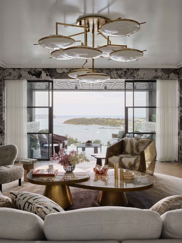 A sophisticated living room with a large sculptural brass-and-glass chandelier, patterned feature wallpaper, floor-to-ceiling glazing, and layered contemporary furnishings. Curved sofas and armchairs surround two organic-shaped marble coffee tables, with a broad harbour and island view visible beyond the windows.