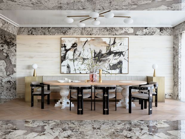 A refined dining room with a long oval timber dining table surrounded by black-framed woven chairs. The room features pale timber wall panelling, dramatic marble-patterned wall and ceiling surfaces, a large abstract artwork, sculptural brass-and-white table lamps, and a contemporary multi-arm ceiling light. The overall composition is luxurious and artful with strong black, white, beige, and natural timber contrasts.